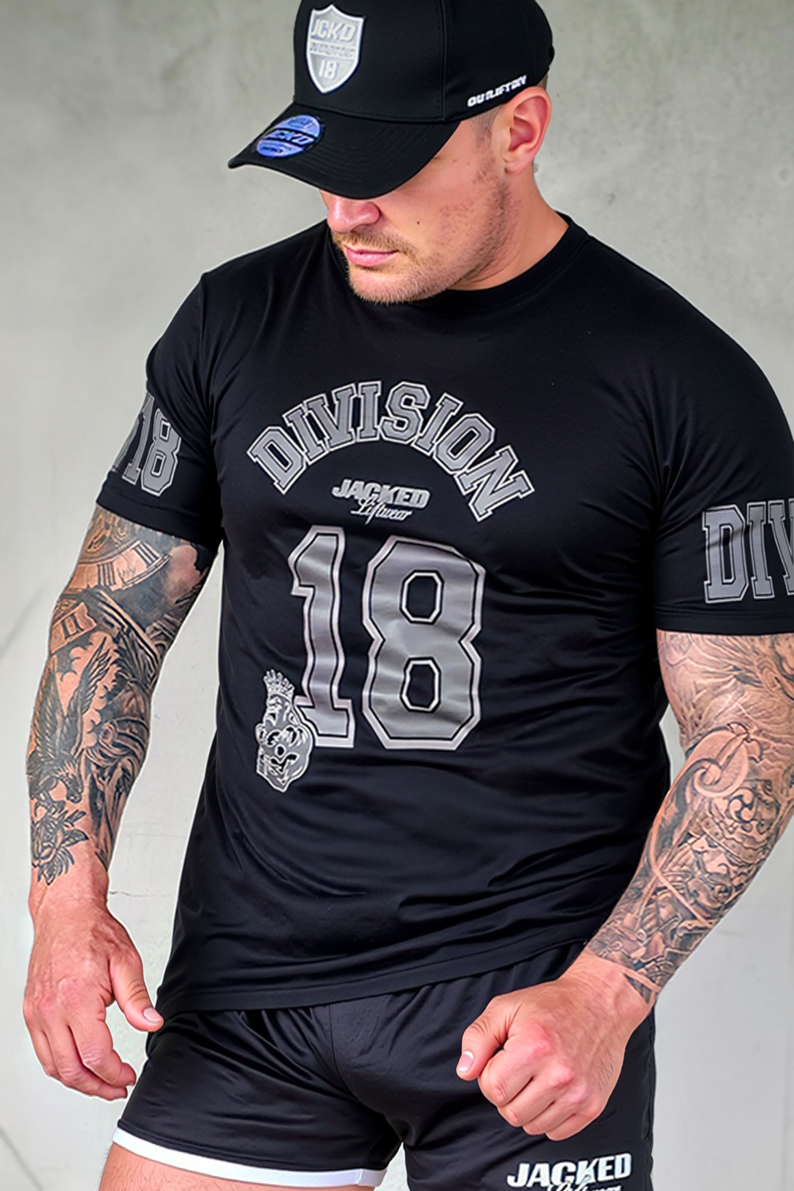 Male fitness model wearing black JCKD Division 18 tee with grey varsity-style graphics and matching JCKD cap, showcasing front print and athletic fit.