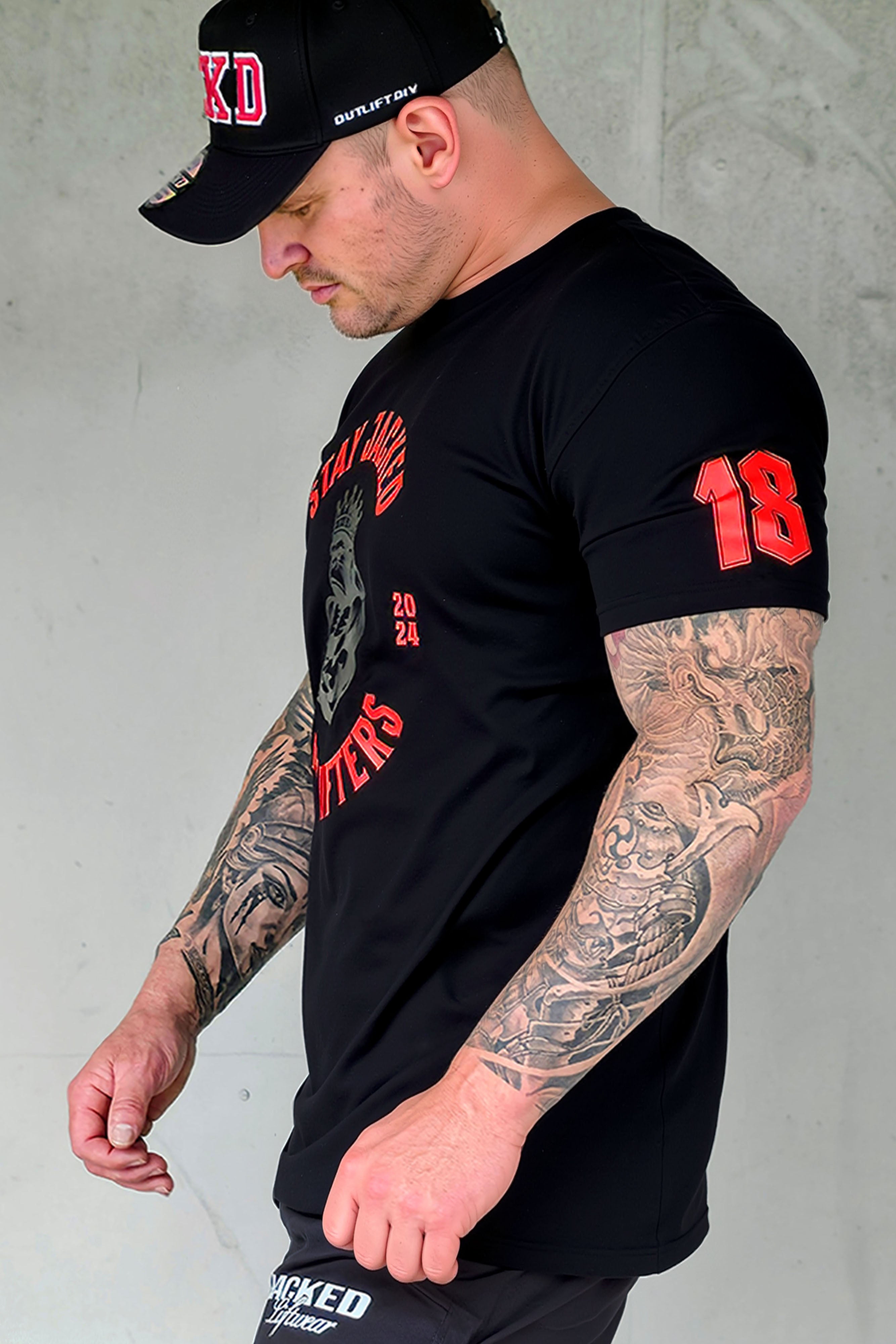 Side view of black Stay Jacked Heavy Lifters tee with red 18 sleeve detail, worn by tattooed male model in JCKD cap.