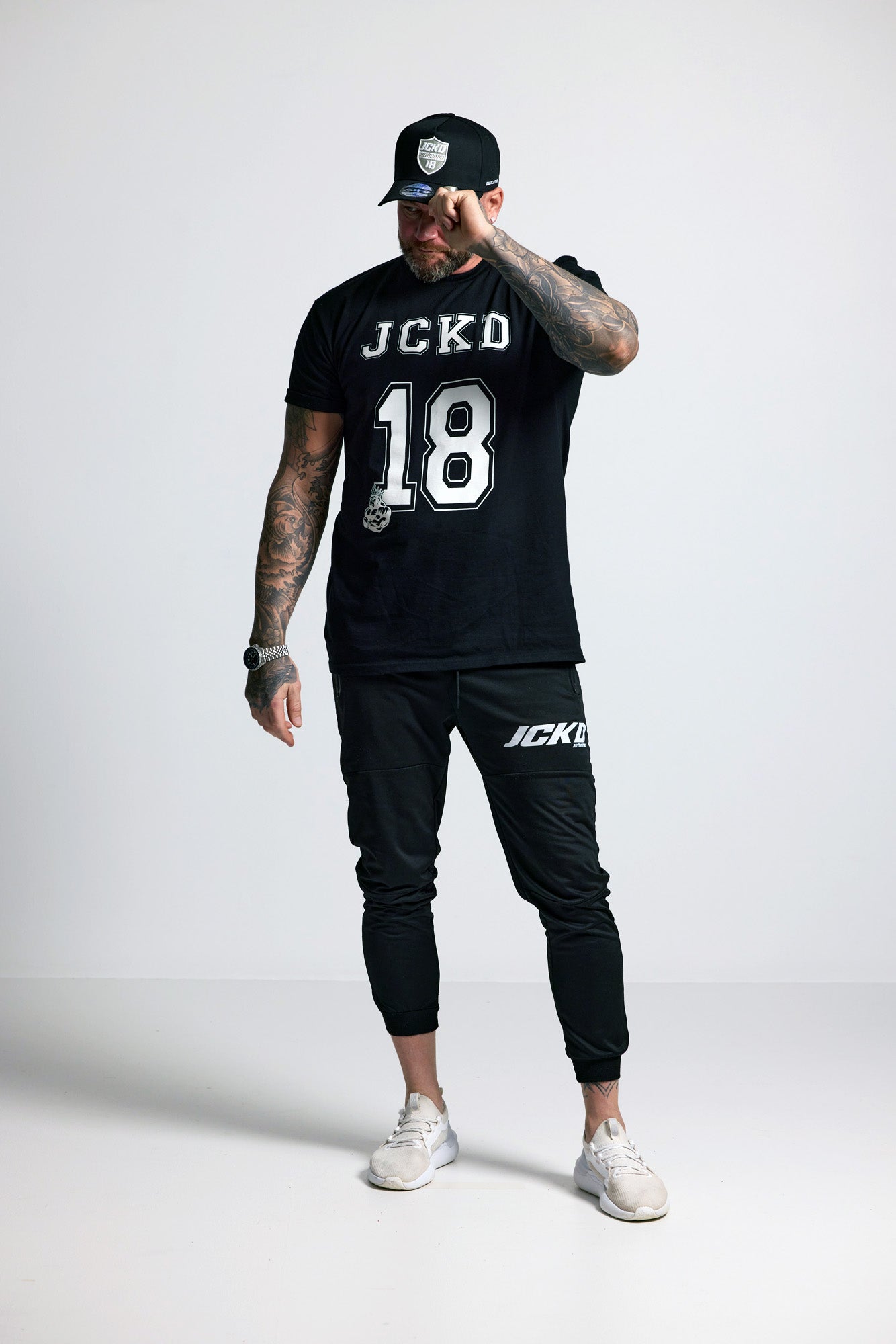 Person wearing a black t-shirt with 'JCKD' and '18', black cap, and black pants on a white background
