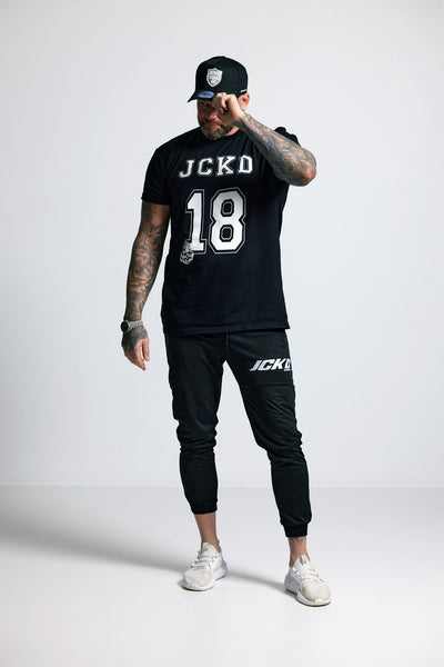 Person wearing a black t-shirt with 'JCKD' and '18', black cap, and black pants on a white background