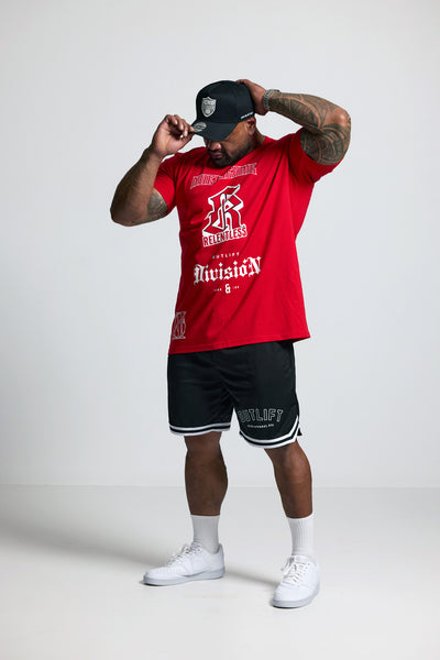 Person wearing a red t-shirt, black shorts, and white sneakers with a cap on a plain background