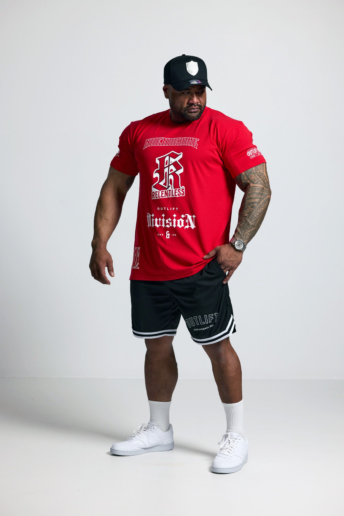 Person wearing a red t-shirt with text and graphics, black shorts, white sneakers, and a black cap on a white background.