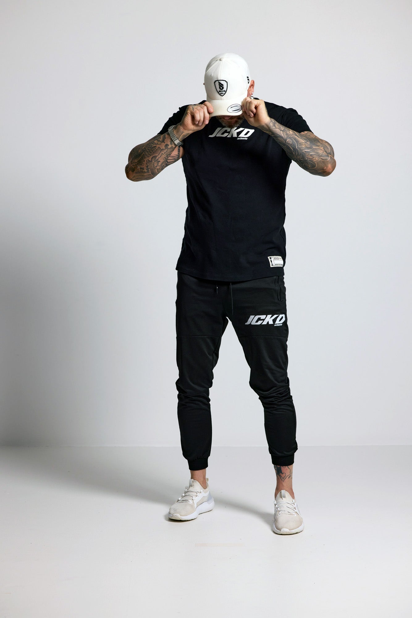 Person wearing black JCKD branded outfit with white cap on a light gray background