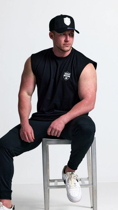 Man sitting on a stool wearing black sleeveless shirt, black pants, and white sneakers with a cap.