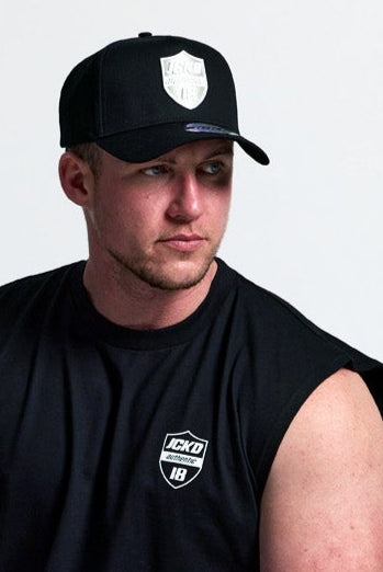Man sitting on a stool wearing black sleeveless shirt, black pants, and white sneakers with a cap.
