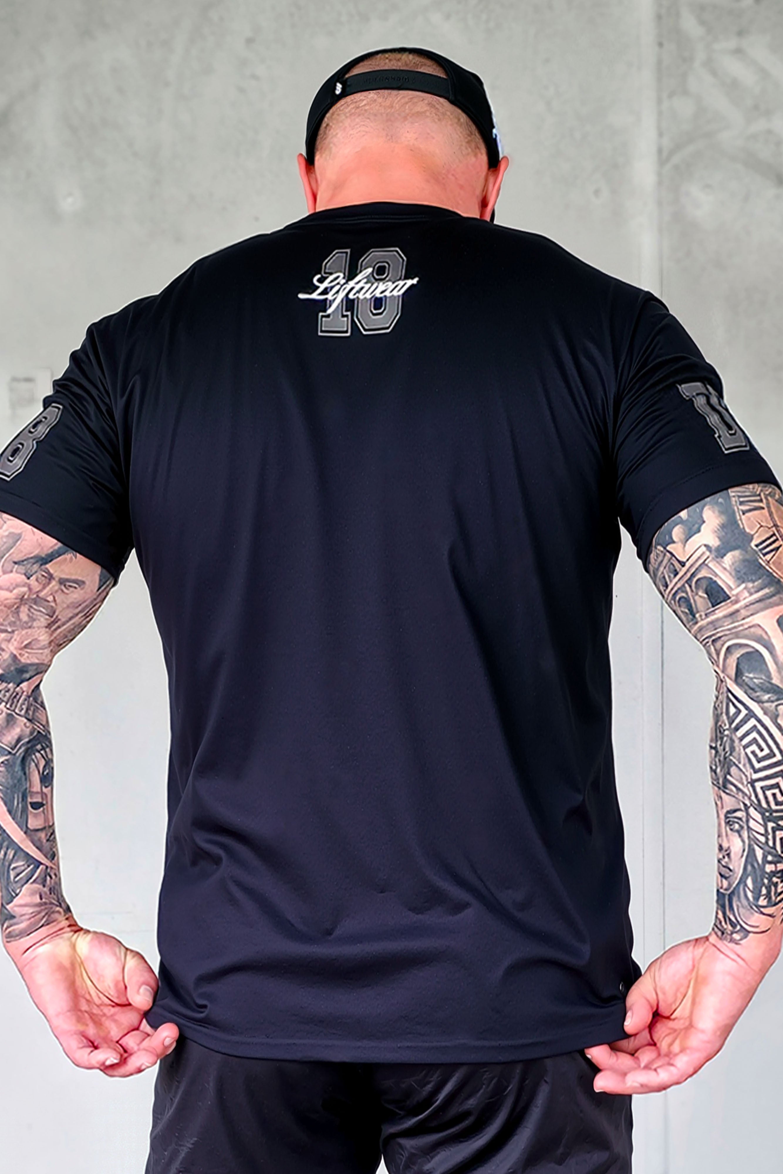 Back view of male model wearing black JCKD Division 18 tee, showcasing the Liftwear 18 upper-back graphic and sleeve detailing.