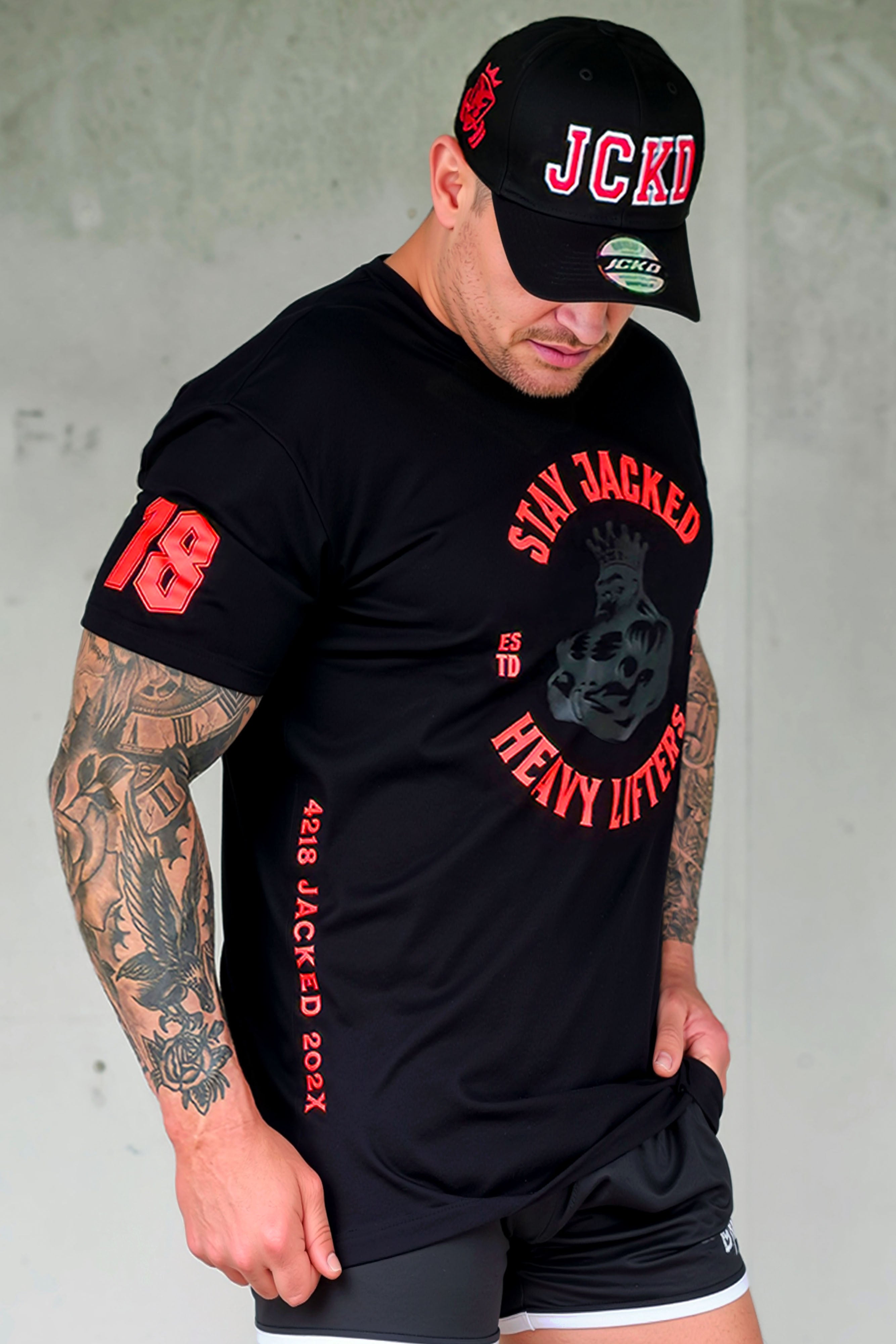 Male fitness model wearing black JCKD cap, Stay Jacked Heavy Lifters tee, and JCKD shorts, showcasing front chest graphic and embroidered hat branding.
