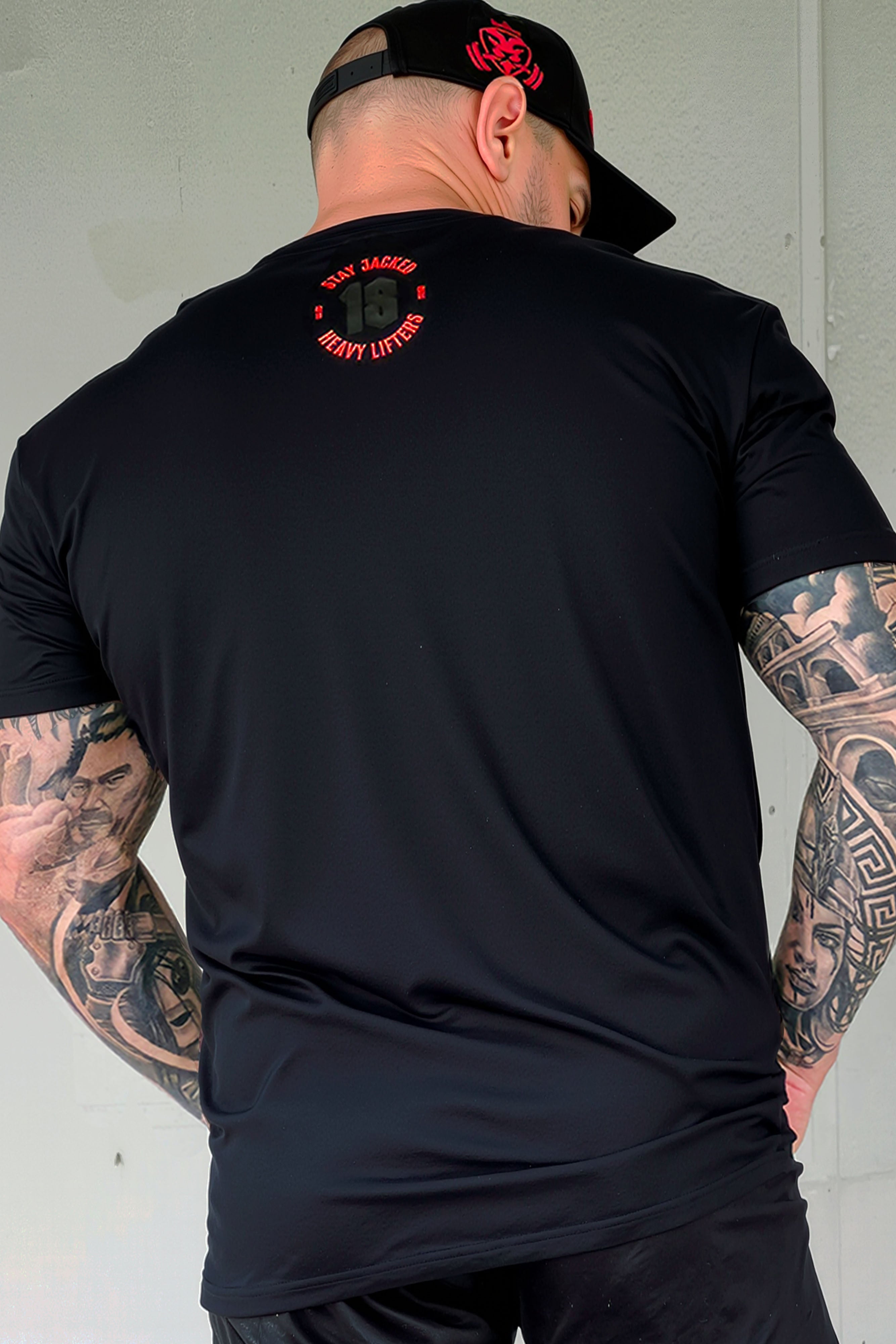Back view of JCKD Stay Jacked Heavy Lifters tee showing rear 18 graphic, worn by a tattooed male model in a black JCKD cap.
