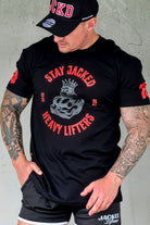 Male fitness model wearing black JCKD cap, Stay Jacked Heavy Lifters tee, and JCKD shorts, showcasing front chest graphic and embroidered hat branding.